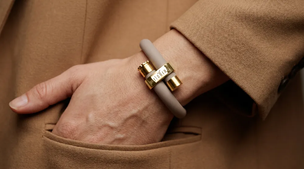 A stone taupe wristraw bracelet with gold accents worn on the wrist, hand resting in a camel coat pocket